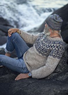 a woman sitting on the rocks by the ocean wearing a sweater and hat with fair hair