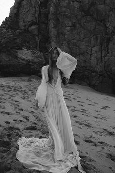 a woman standing on top of a sandy beach next to a rocky cliff and wearing a white dress