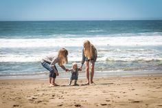 two women and a child on the beach with waves in the background, one holding her hand