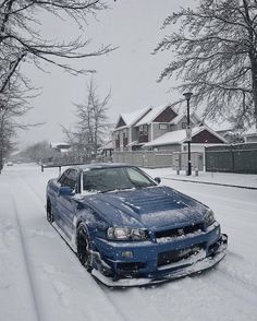 a blue car parked in the middle of a snow covered street next to trees and houses