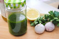 a jar filled with green liquid next to garlic, lemon and parsley on a cutting board