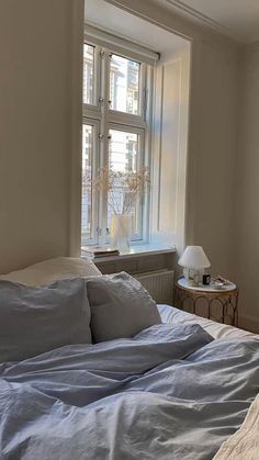 an unmade bed sitting in front of a window next to a nightstand with a lamp on it