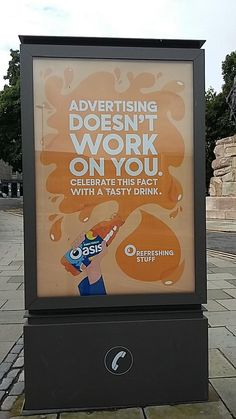 an advertising sign on the side of a road near a building and water fountain with trees in the background