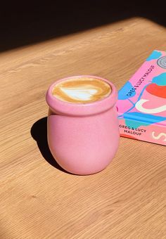 a cup of coffee sitting on top of a wooden table next to a pink book