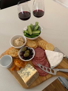 a wooden cutting board topped with cheese, crackers and meats next to two glasses of wine