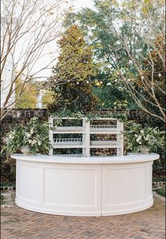 a white bench sitting on top of a brick floor next to trees and shrubbery