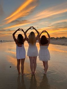 three young women standing on the beach making heart shapes with their hands while the sun sets