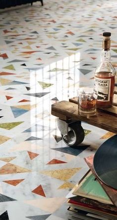 a wooden table topped with a bottle of alcohol on top of a floor covered in colorful tiles