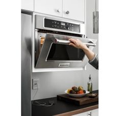 a person opening the door to a stainless steel microwave oven in a kitchen with white cabinets