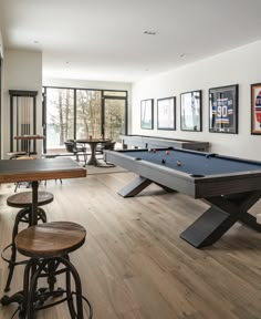 a pool table and some stools are in a room with wood flooring, windows, and pictures on the wall