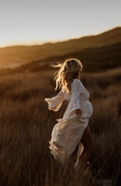 a woman in a white dress is running through the grass at sunset with her hair blowing in the wind