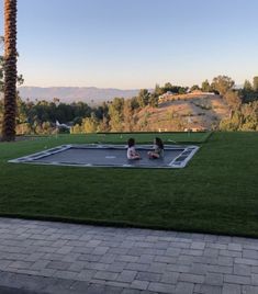 two people sitting on the ground in front of a swimming pool with mountains in the background