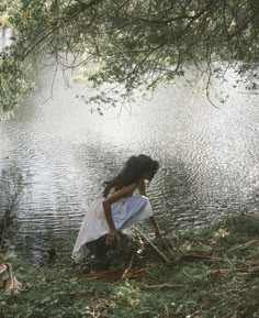 a woman kneeling down next to a body of water with trees in front of her