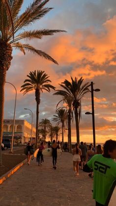 a group of people walking down a sidewalk with surfboards under palm trees at sunset