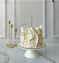 a white cake sitting on top of a table next to two gold candles and a wall