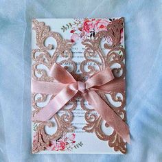 a pink and gold wedding card with a bow on it's side, sitting on top of a blue cloth