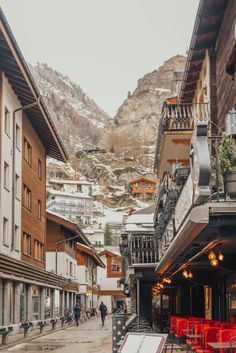 an old town with mountains in the background and red chairs on the sidewalk, people walking down the street