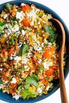 a blue bowl filled with vegetables and feta cheese next to a wooden spoon on a white surface