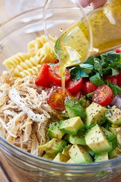 someone pouring dressing into a bowl filled with pasta, avocado, tomatoes and cucumbers