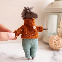 a hand holding a tiny knitted doll in front of a white table with yarn balls