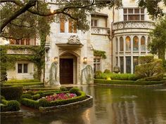 a large house with lots of trees and flowers in the front yard, next to a pond