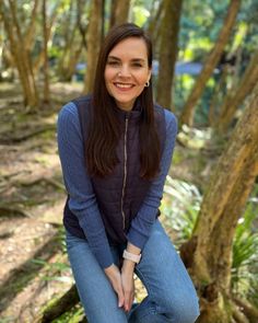 a woman sitting in the woods with her hands on her knees and smiling at the camera