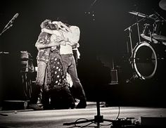 two people are hugging on stage in front of microphones and drum set behind them