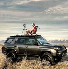 a man sitting on top of a vehicle in the middle of a field with mountains behind him