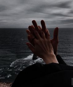 a person holding their hands up in the air near the ocean on a cloudy day