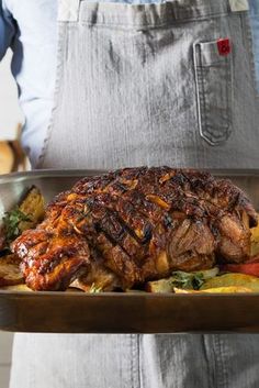 a person in an apron holding a roasting pan filled with meat and vegetables