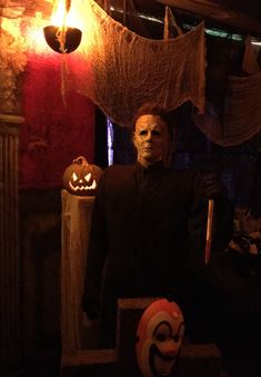 a creepy man in a black suit holding a broom and wearing a mask with two jack - o'- lanterns hanging from the ceiling