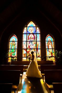 a bride and groom standing in front of stained glass windows at the end of a church aisle