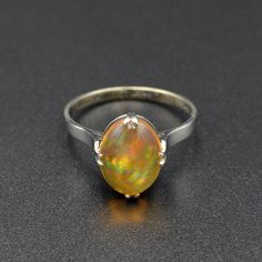 "A rainbow is swimming in this gorgeous antique opal ring! A beautifully proportioned opal cabochon is centered with cardinal prongs (north, south, west and east) in an 18k white gold setting. The opal features a transparent orange body, typical of Mexican jelly opals, with a \"contra luz\" color flash seen when the gem interacts with light. The color play is astounding, with every color of the rainbow ignited within the body of the opal, having painterly brush-stroke like patterns. This ring hails from the 1920s and has been tenderly well-cared for in its lifetime. It is in wonderful condition and ready for a new owner. This ring could be a perfect alternative engagement ring, gift for someone with an October birthstone or simply a present for an antique jewelry lover.   Opal is a fascina Ethiopian Opal Round Ring Fine Jewelry, Antique Opal Ring, Jelly Opal, Color Play, Etsy Gold Ring, Alternative Engagement Rings, North South, October Birthstone, Solitaire Rings