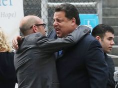 two men hug each other as they stand in front of a fence with people behind them