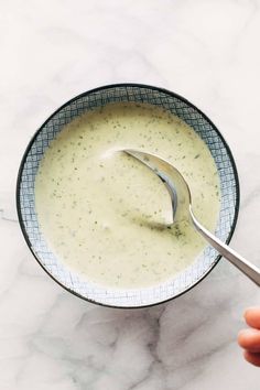 a hand holding a spoon over a bowl of broccoli soup on a marble table