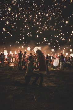 many people are standing in the grass with lanterns floating above them at night time,