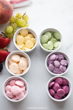 six bowls filled with different colored marshmallows next to some fruit on a table