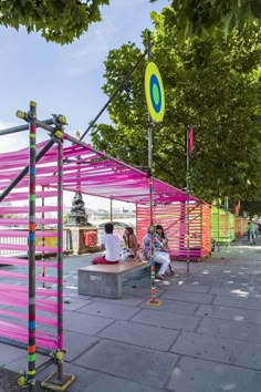 several people sitting on benches under pink and yellow awnings at an outdoor park