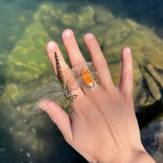 a hand holding a tiny orange and black dragonfly on it's thumb next to some water