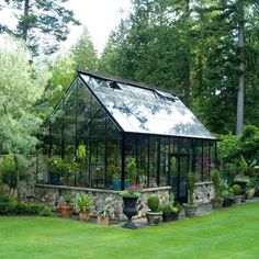 a small glass house in the middle of a green yard with potted plants and trees