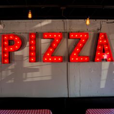 the word pizza is lit up in red letters on a white wall behind two tables with red checkered tablecloths