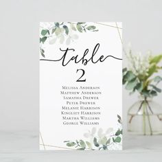 a white table number card with greenery on it and a vase filled with flowers