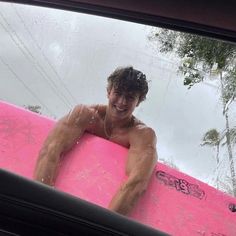 a young man holding a pink surfboard in the back seat of a car with trees and water behind him