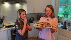 two girls are standing in the kitchen and one is holding a tray with cookies on it