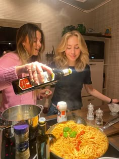 two women in the kitchen pouring sauce over spaghetti noodles on top of a skillet