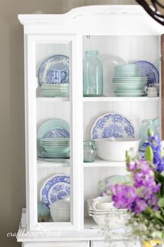 a white china cabinet with blue and white dishes on the top, purple flowers in vase next to it