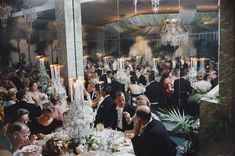 a large group of people sitting at tables in a room with chandeliers hanging from the ceiling