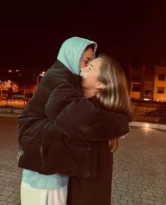 two people hugging each other on a brick walkway at night time with buildings in the background