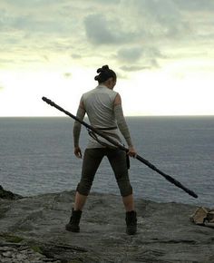 a woman standing on top of a rock next to the ocean holding two giant swords