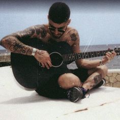 a man with tattoos playing an acoustic guitar on top of a roof next to the ocean